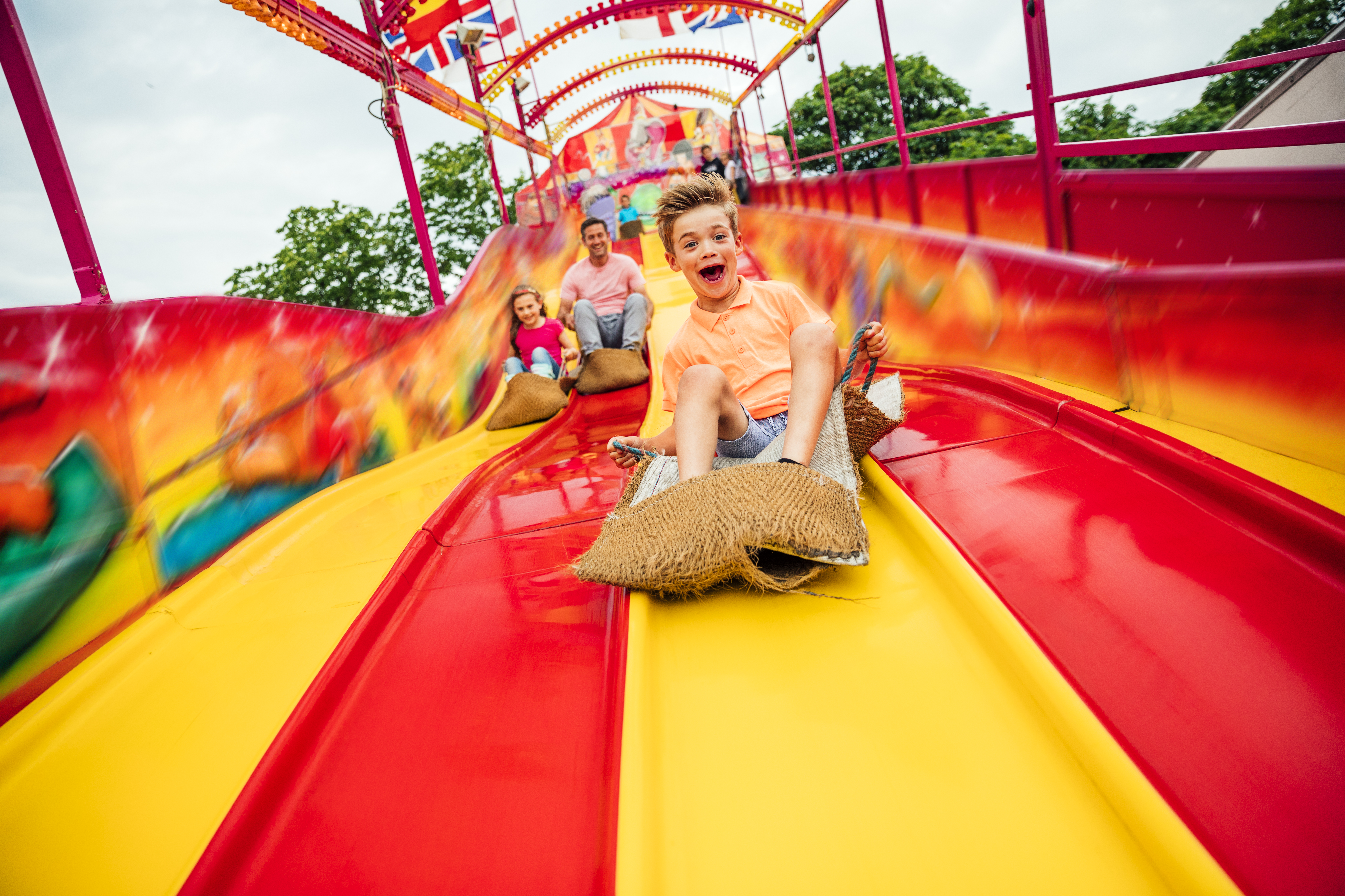 little boy having fun sliding down a yellow and red slide while sitting in a burlap sack